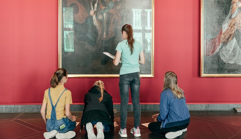 Das Foto zeigt vier Schülerinnen von hinten, die, mit Stift und Papier ausgestattet, das große Porträt eines sächsischen Herrschers aus der Barockzeit betrachten. Rechts von diesem Gemälde hängt ein großes Porträt einer weiblichen Person aus dieser Zeit.