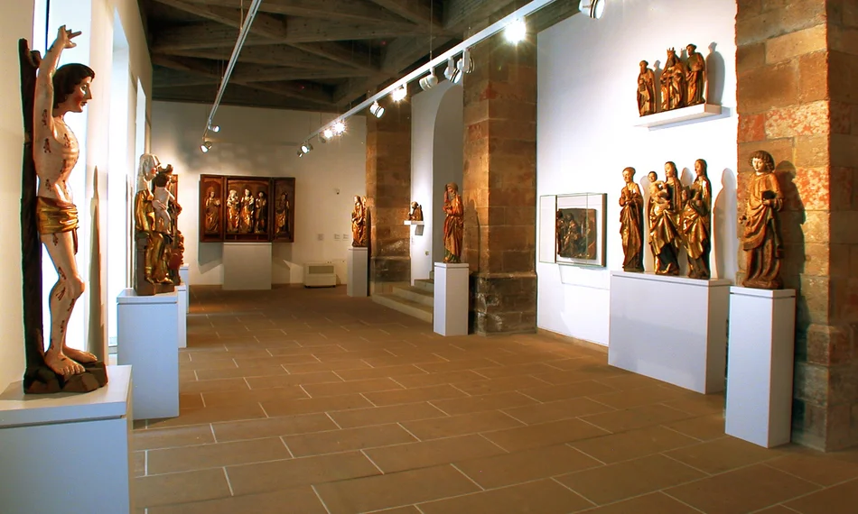 Blick in die Dauerausstellung „Gotische Skulptur in Sachsen“ Das Foto zeigt einen Blick in die Dauerausstellung „Gotische Skulptur in Sachsen“, die sich in den rekonstruierten historischen Klosterräumen des Schloßbergmuseums befindet. Zu sehen sind mehrere Werke sakraler Kunst, hauptsächlich mittelalterliche Heiligenstatuen, aber auch ein Flügelaltar mit Bildern biblischer Szenen.