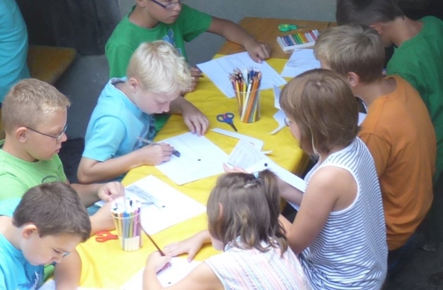 Kinder sitzen an einem Tisch und arbeiten mit Buntstiften, Schere und Leim auf Papier.