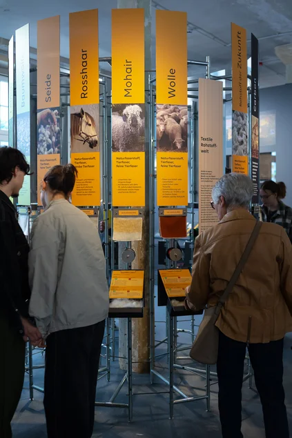 Turm der Tuche An einem kreisförmig angeordneten Ausstellungselement, an dem Text- und Bildtafeln angebracht sind, stehen mehrere Besucherinnen und Besucher.