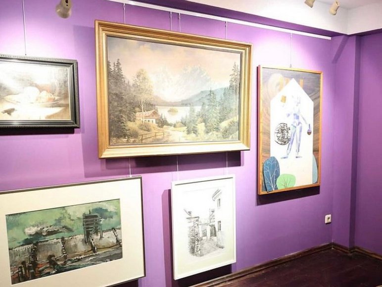 An einer Ausstellungswand in Violett sind fünf im Salon-Stil gehängte Bilder angeordnet..