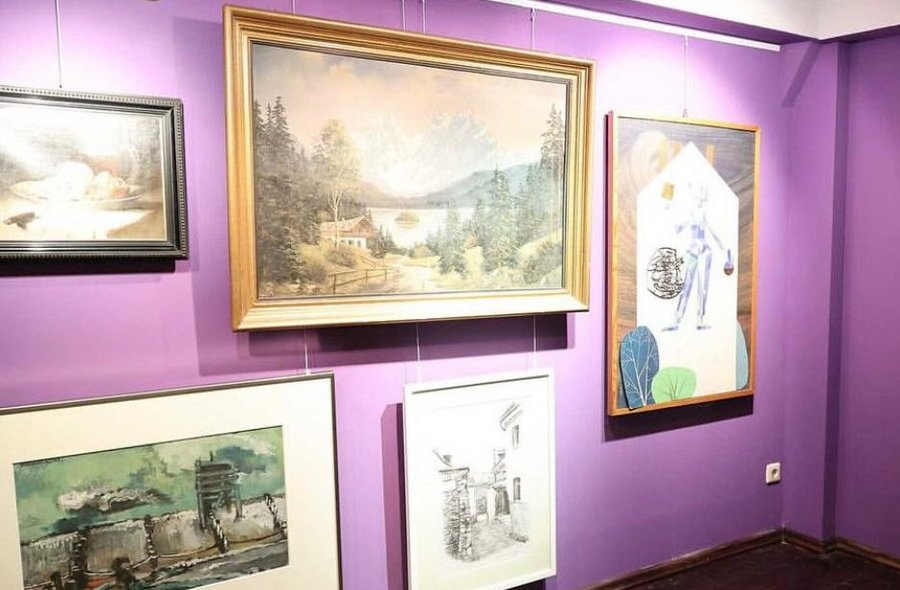 An einer Ausstellungswand in Violett sind fünf im Salon-Stil gehängte Bilder angeordnet..