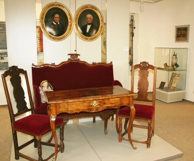 Das Foto zeigt einen Biedermeiertisch mit zwei weinrot gepolsterten Stühlen und einem weinrot gepolsterten Sitzmöbel für zwei Personen, im Hintergrund zwei in ovale Goldrahmen gefasste historische Porträts und in der Ecke eine kleine Vitrine mit diversen Exponaten zur Stadtgeschichte.