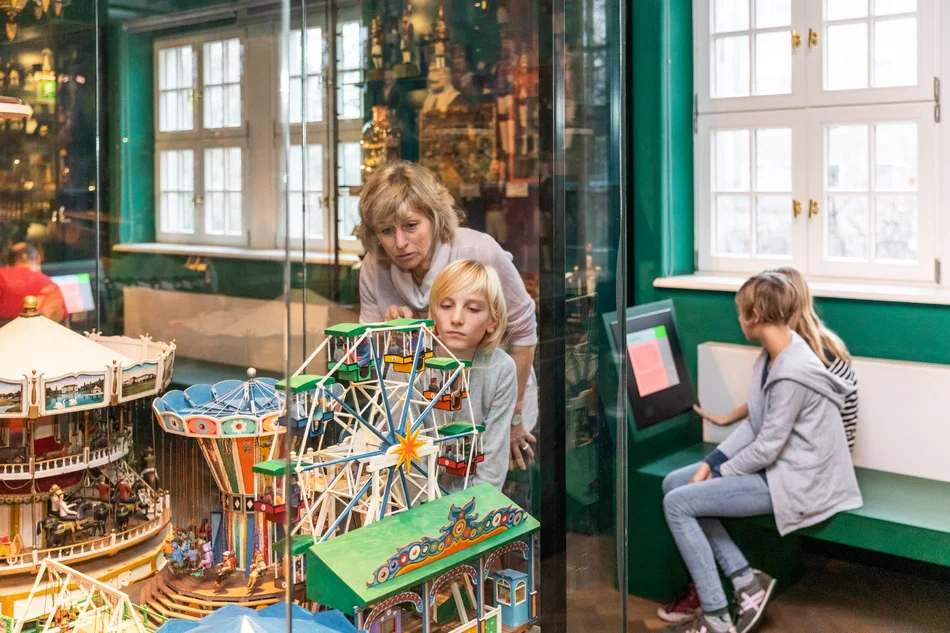 Eine Frau und ein Kind blicken gemeinsam in eine mit Spielzeug-Karussells bestückte Vitrine. Im Hintergrund sitzt eine Person an einer Medienstation.