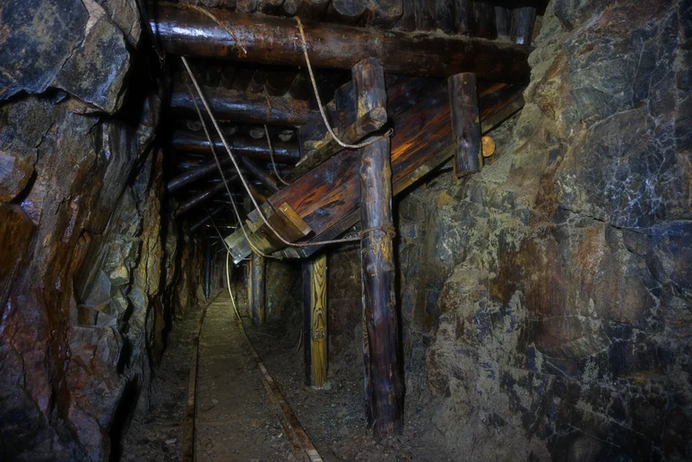 Ein enger Gang durch einen Stollen, am Boden alte Gleise. An der rechten Seite und der Decke finden sich Balken und Stämme in einer Holzkonstruktion, einige Bretter sind mit Seilen gesichert.