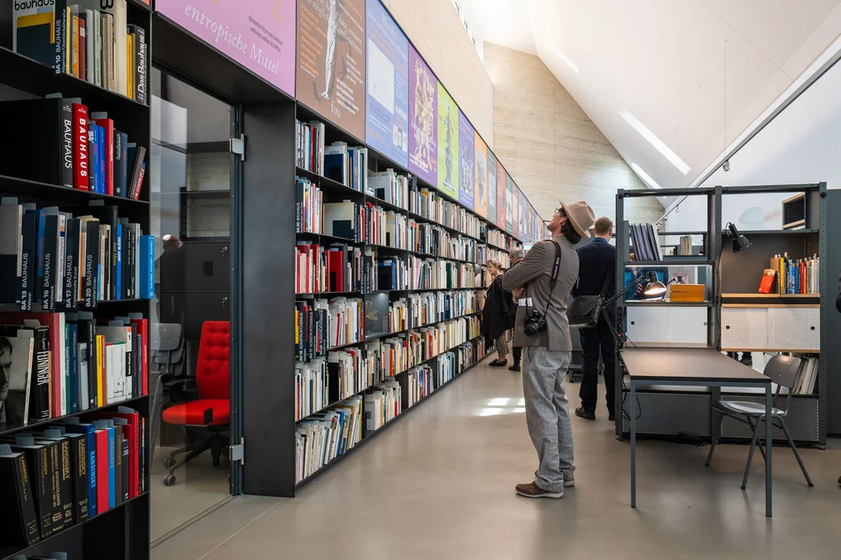 In einem bibliothek-artigen Raum betrachtet ein Mann mit Hut das breite Bücherregal. Hinter ihm befindet sich ein Arbeitsplatz mit Tisch und Regal.