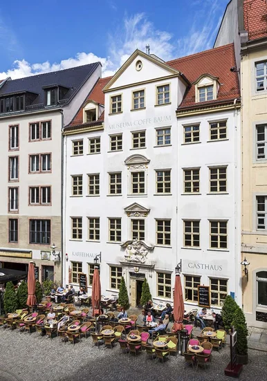 Außenansicht eines fünfstöckigen Barockhauses in einer historischen Häuserzeile. Direkt vor dem Haus stehen Café-Tische und -Stühle.