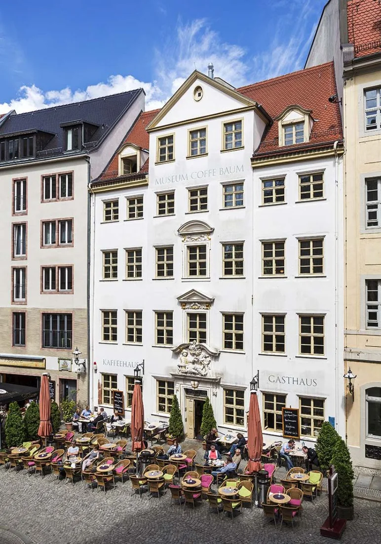 Außenansicht eines fünfstöckigen Barockhauses in einer historischen Häuserzeile. Direkt vor dem Haus stehen Café-Tische und -Stühle.