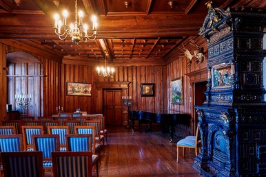 Kaminzimmer im Schloss Hinterglauchau In einem holzvertäfelten historischen Raum mit Kronleuchtern stehen ein reich verzierter alter Kachelofen, ein Flügel und mehrere Stuhlreihen.