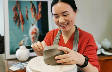 Eine Porzellankünstlerin arbeitet an einem Rohling, im Hintergrund sind eine schlichte weiße Vase sowie ein Bild mit dem Foto einer bemalten Porzellanvase zu sehen.
