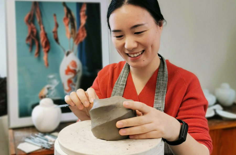 Eine Porzellankünstlerin arbeitet an einem Rohling, im Hintergrund sind eine schlichte weiße Vase sowie ein Bild mit dem Foto einer bemalten Porzellanvase zu sehen.
