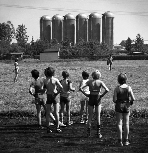 Schwarzweißfotografie einer sommerlichen Szene, in der mit dem Rücken zum Fotografen stehende, in Sportsachen gekleidete Kinder auf einer Wiese stehen und in Richtung zweier Erwachsener blicken, im Hintergrund stehen markant sechs Silos.
