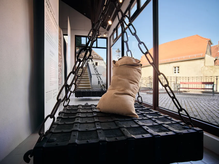 Das Foto zeigt eine monumentale gusseiserne Balkenwaage – die in Oschatz ausgestellte Salzwaage aus dem Jahr 1686. Auf einer der Waagflächen steht ein prall gefüllter weißer Sack.