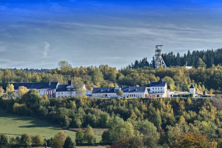 Sommerliche Außenansicht des inmitten einer bewaldeten Fläche befindlichen Gebäudekomplexes mit dem Förderturm im Hintergrund