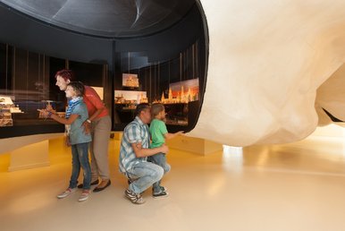 Foto einer vierköpfigen Familie in einem Ausstellungsraum, die Eltern schauen sich mit ihren Kindern Volkskunst-Exponate in einer großen, geschwungenen Vitrine an.