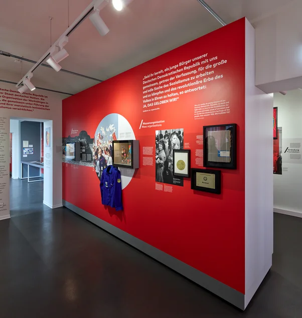 Das Foto zeigt eine große Ausstellungswand. Vor einem roten Hintergrund sind Schwarzweiß-Fotos von jungen Leuten bei der Jugendweihe in der DDR zu sehen und ein blaues FDJ-Hemd. Daneben steht ein Zitat. Es lautet: Zitat "Seid ihr bereit, als junge Bürger unserer Deutschen Demokratischen Republik mit uns gemeinsam, getreu der Verfassung, für die große und edle Sache des Sozialismus zu arbeiten und zu kämpfen und das revolutionäre Erbe des Volkes in Ehren zu halten, so antwortet: Ja, das geloben wir!" Zitat Ende.