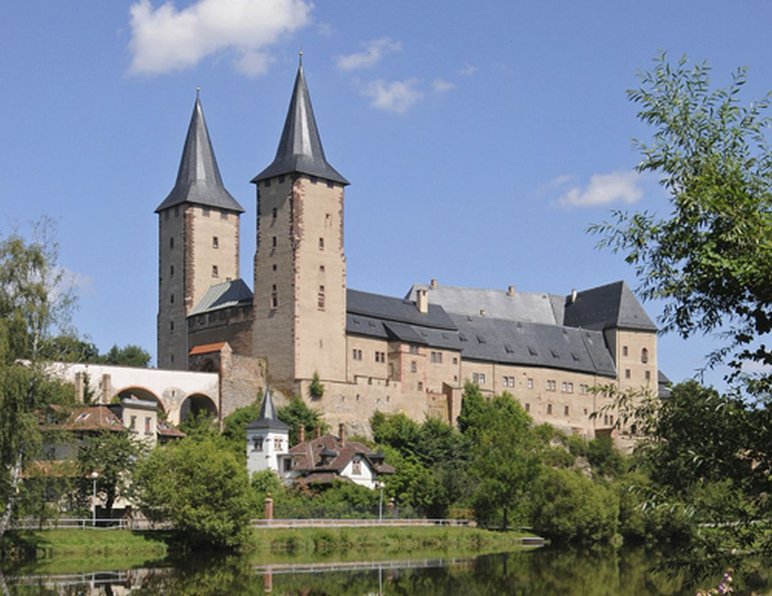 Sommerliche Außenansicht des Schlosses, welches in seiner Anmutung eher einer Burg ähnelt. Prominent stechen zwei Spitztürme hervor.