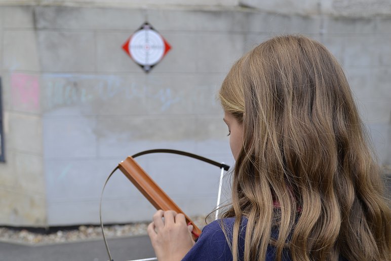 Ein von hinten fotografiertes Mädchen spannt eine Armbrust; auf der Mauer im Hintergrund ist eine Zielscheibe.
