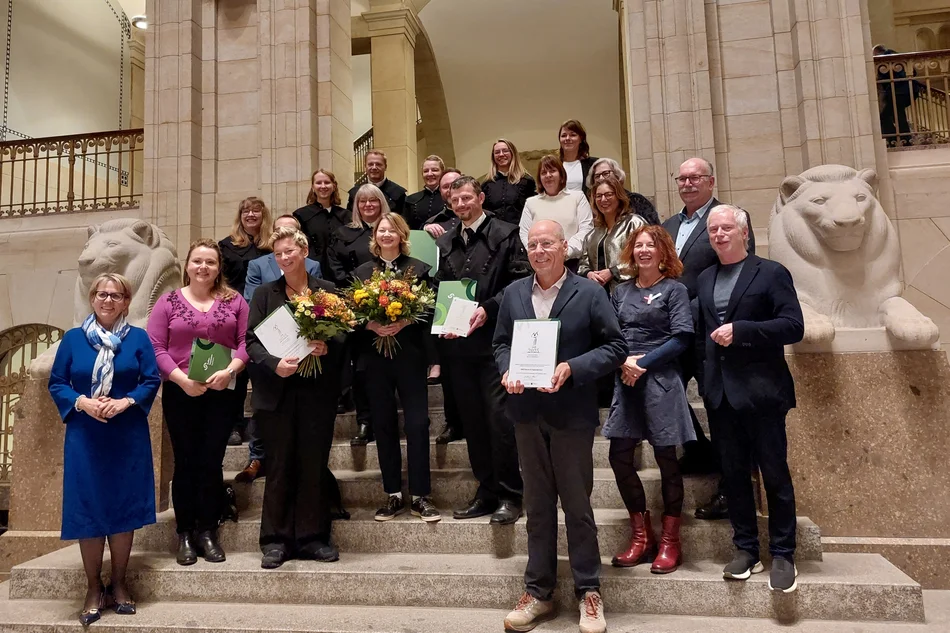Festlich gekleidete Personen stehen auf den Stufen einer repräsentativen Säulenhalle, einige mit Blumensträußen oder Urkunden in den Händen
