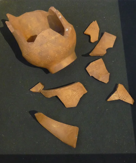 Ein beschädigtes Ton-Gefäß und mehrere Scherben liegen auf schwarzem Untergrund.