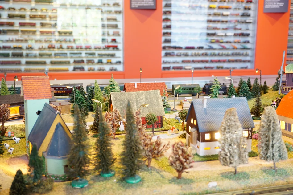 Das Foto zeigt eine Detailaufnahme der Modellbahnsammlung Ziemert. Zu sehen ist ein Ausschnitt einer Modelleisenbahn-Landschaft mit Häuschen und Bäumchen. Die Wand im Hintergrund ist mit Vitrinen mit zahlreichen Modelleisenbahnen bestückt.