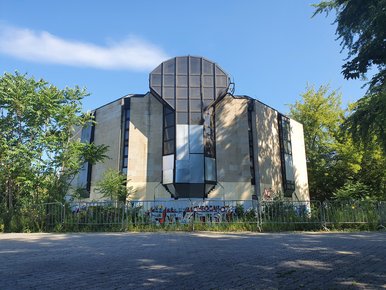 Der ehemalige Bowlingtreff Hinter einer Metallabsperrung ragt, umwuchert von ungemähtem Gras und Bäumen, ein markant gestaltetes Beton-Gebäude auf.