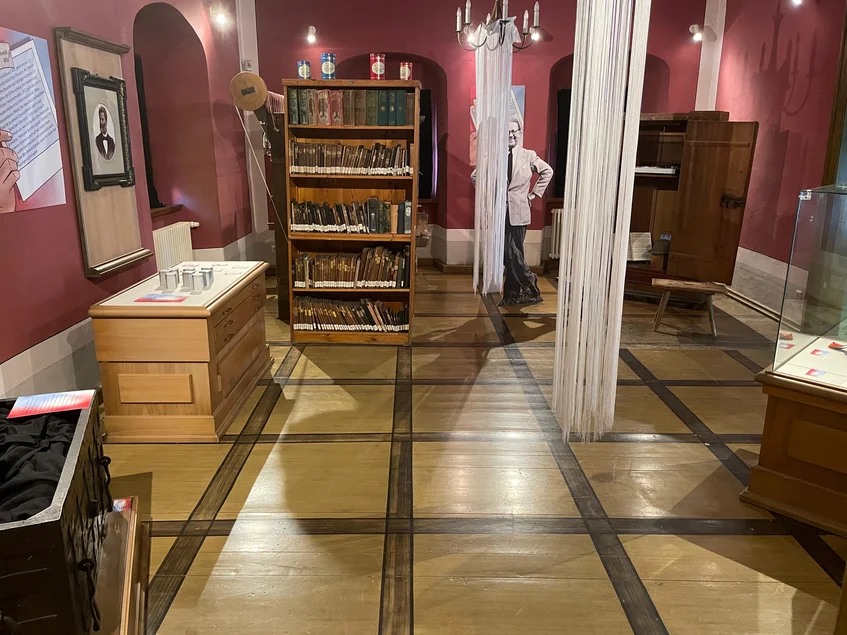 In einem historisch anmutenden Raum mit Holzfußboden stehen mehrere geöffnete Schränke und Truhen sowie eine Vitrine und ein Bücherregal. Dazwischen steht das lebensgroße Bild eines älteren Mannes.