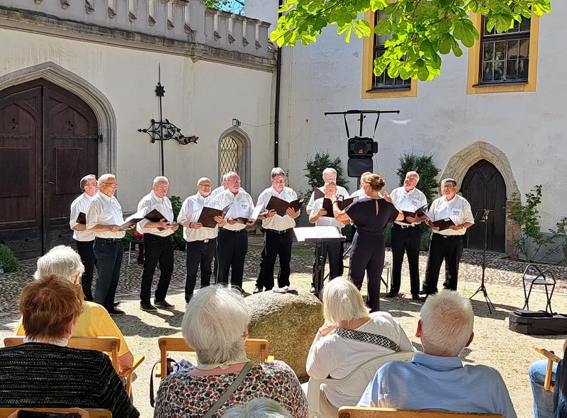 Ein kleiner Männerchor singt bei schönem Wetter im Freien. Das Publikum sitzt auf Stühlen unter einem Baum.