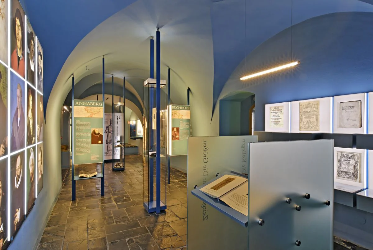 Blick in die Dauerausstellung In einem historischen Gewölbe hängen hinterleuchtete Ausstellungselemente, zentral steht ein Tisch mit einem geöffneten Buch.