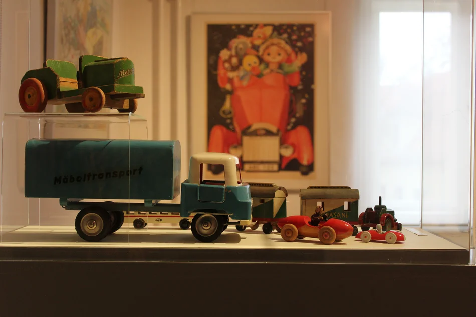 In einer Vitrine stehen unterschiedliche historische Spielzeugfahrzeuge aus Holz, unter anderem ein Möbeltransporter und ein Traktor.