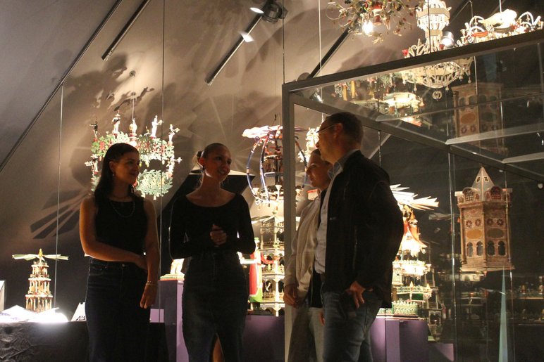Vier Personen stehen in einem Ausstellungsraum, in dem eine Vielzahl erzgebirgischer Weihnachtspyramiden und andere Schnitzkunst präsentiert werden.
