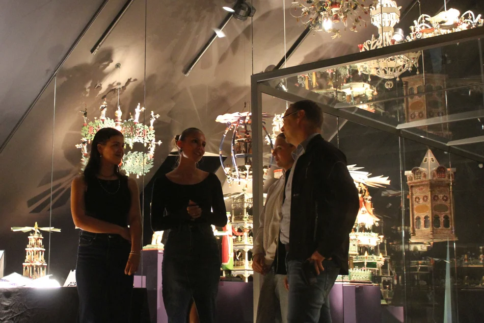 Vier Personen stehen in einem Ausstellungsraum, in dem eine Vielzahl erzgebirgischer Weihnachtspyramiden und andere Schnitzkunst präsentiert werden.