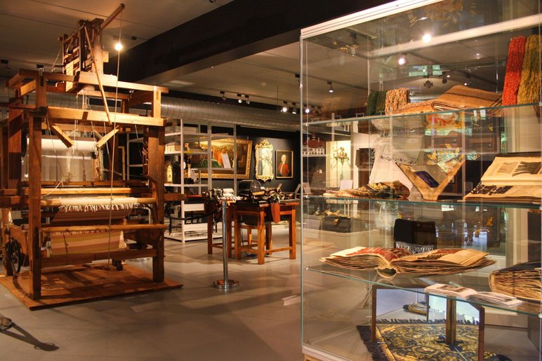 Ein Handwebstuhl, Textilmaschinen und historische Stoffe sind im Schaudepot neben Gemälden und anderen stadtgeschichtlichen Objekten in Regalen zu sehen.