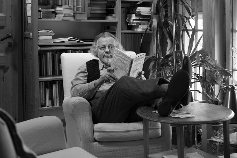 Schwarzweißfotografie von Thomas Rosenlöcher, der, gemütlich in einem Sessel sitzend, von seinem Buch aufblickt