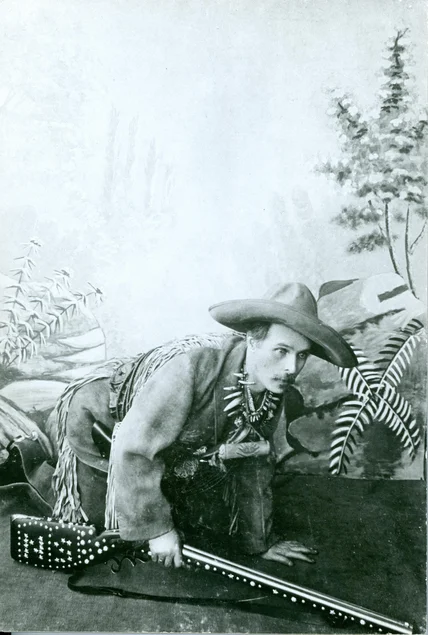 Ein als Cowboy verkleideter Mann auf allen Vieren, sich scheinbar anschleichend, in der rechten Hand ein Gewehr haltend