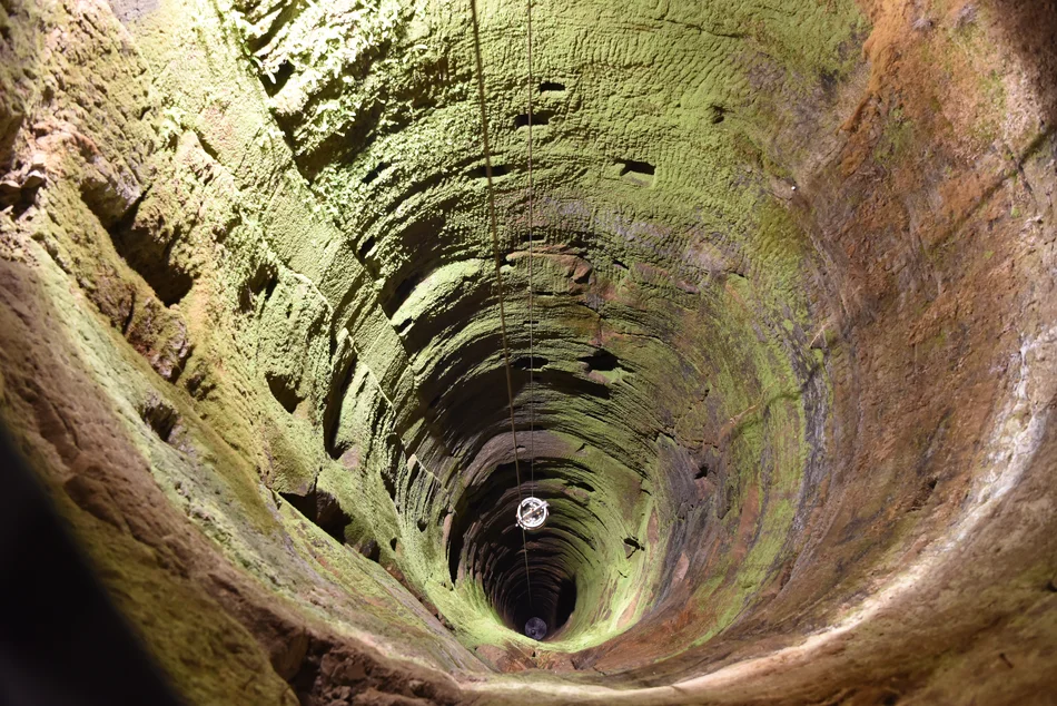 Blick in den 152 Meter tiefen Brunnenschacht Beleuchteter Brunnenschacht der Festung Königstein. Ein gefülltes Wasserfass hängt an einem Seil und wird nach oben gezogen.