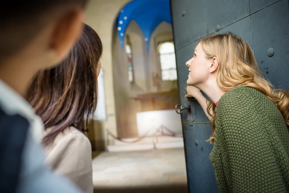 Sonderführung zum Tag des offenen Denkmals 2025 Drei Personen blicken durch eine halb geöffnete, alte eiserne Schlosstür in einen mit Rundbögen versehenen historischen Raum.