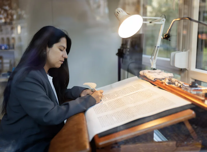 Eine an einem Pult sitzende Frau beschreibt mit einem Federkiel ein großes Papier in kleiner, gleichmäßiger Schrift.