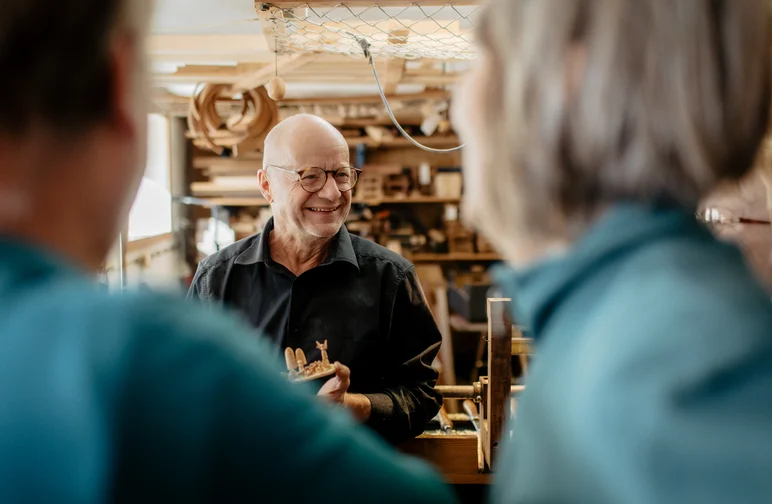 Ein mit Holz arbeitender Kunsthandwerker präsentiert zwei Besuchenden seine Arbeit.