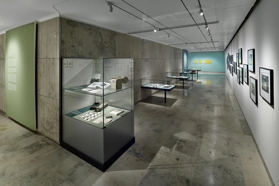 Auf dem Foto ist der Außenbereich des Buchmuseums abgebildet, sozusagen die Galerie, in deren Innerem sich wie ein Kubus die Schatzkammer befindet. Zu sehen ist links die grau-braune Travertin-Wand, die die Schatzkammer umgibt, davor befinden sich vier Flach- und eine Hochvitrine. Rechts an der Wand sind gerahmte Fotografien zu erkennen.