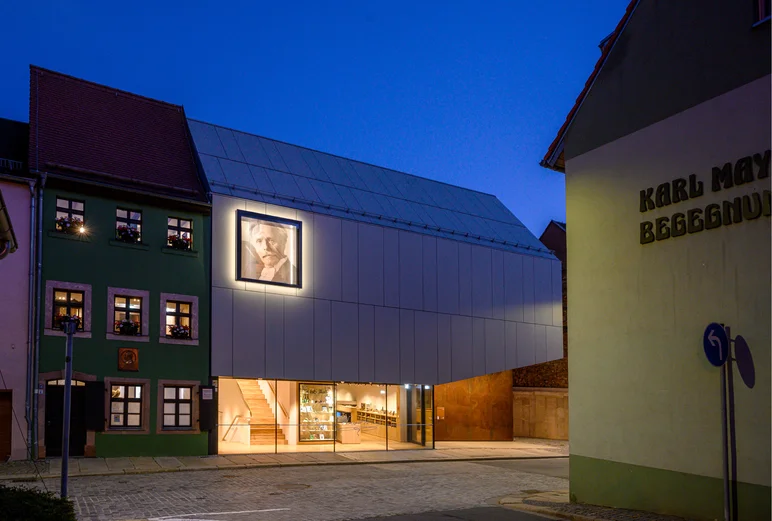 Das Foto zeigt eine Außenansicht des Karl-May-Hauses und des Museumsdepots bei Nacht. Der Eingangsbereichs des Museumsneubaus aus hell erleuchtet.