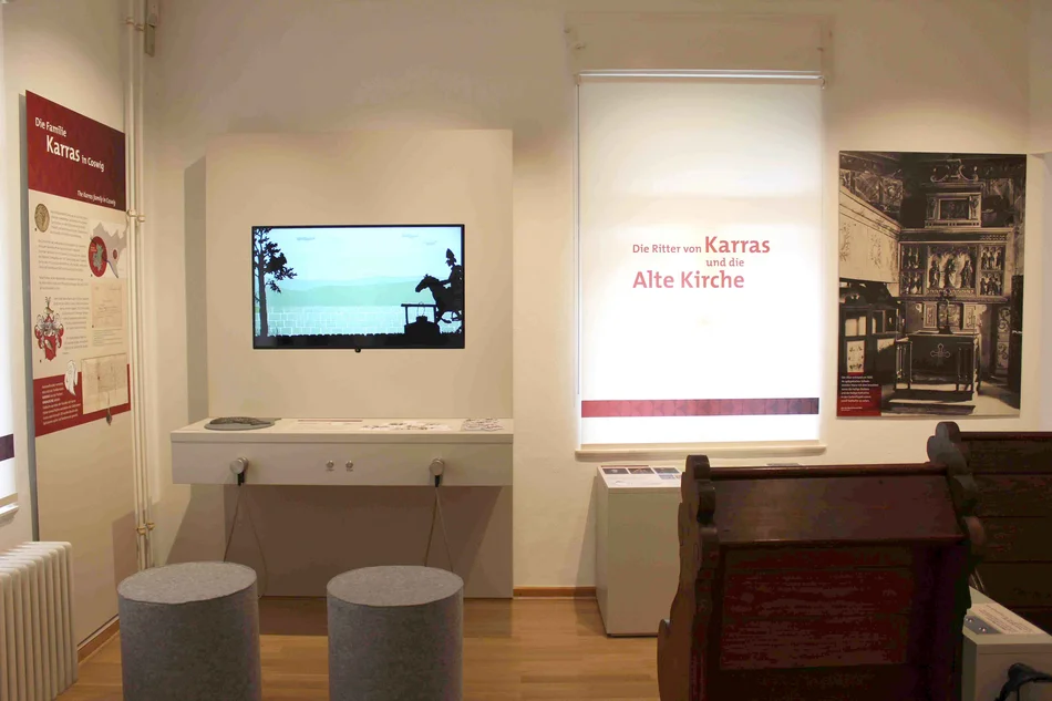 Einblick in den Raum zu den Karras und der Alten Kirche Im Raum zu sehen ist historisches Kirchengestühl, zentraler Blickpunkt des Fotos ist eine unterfahrbare Medienstation mit zwei Sitzhockern davor.