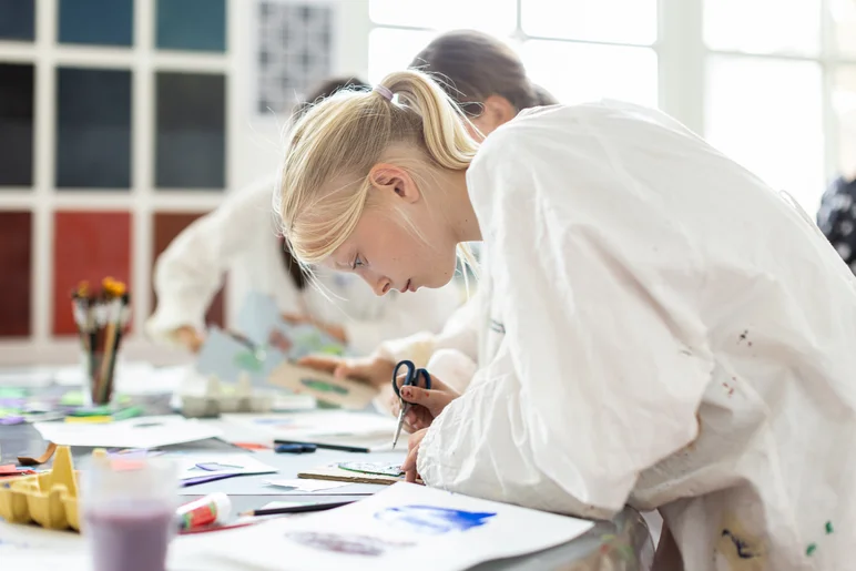 Das Foto zeigt eine Schülerin bei einem Vermittlungsangebot in der Gemäldegalerie Alte Meister der Staatlichen Kunstsammlungen Dresden. Sie trägt einen weißen Kittel und ist, Schere in der Hand, über einen großen Arbeitstisch gebeugt. Auf dem Tisch befindet sich eine Vielzahl verschiedener Materialien sowie ein Becher mit Pinseln.