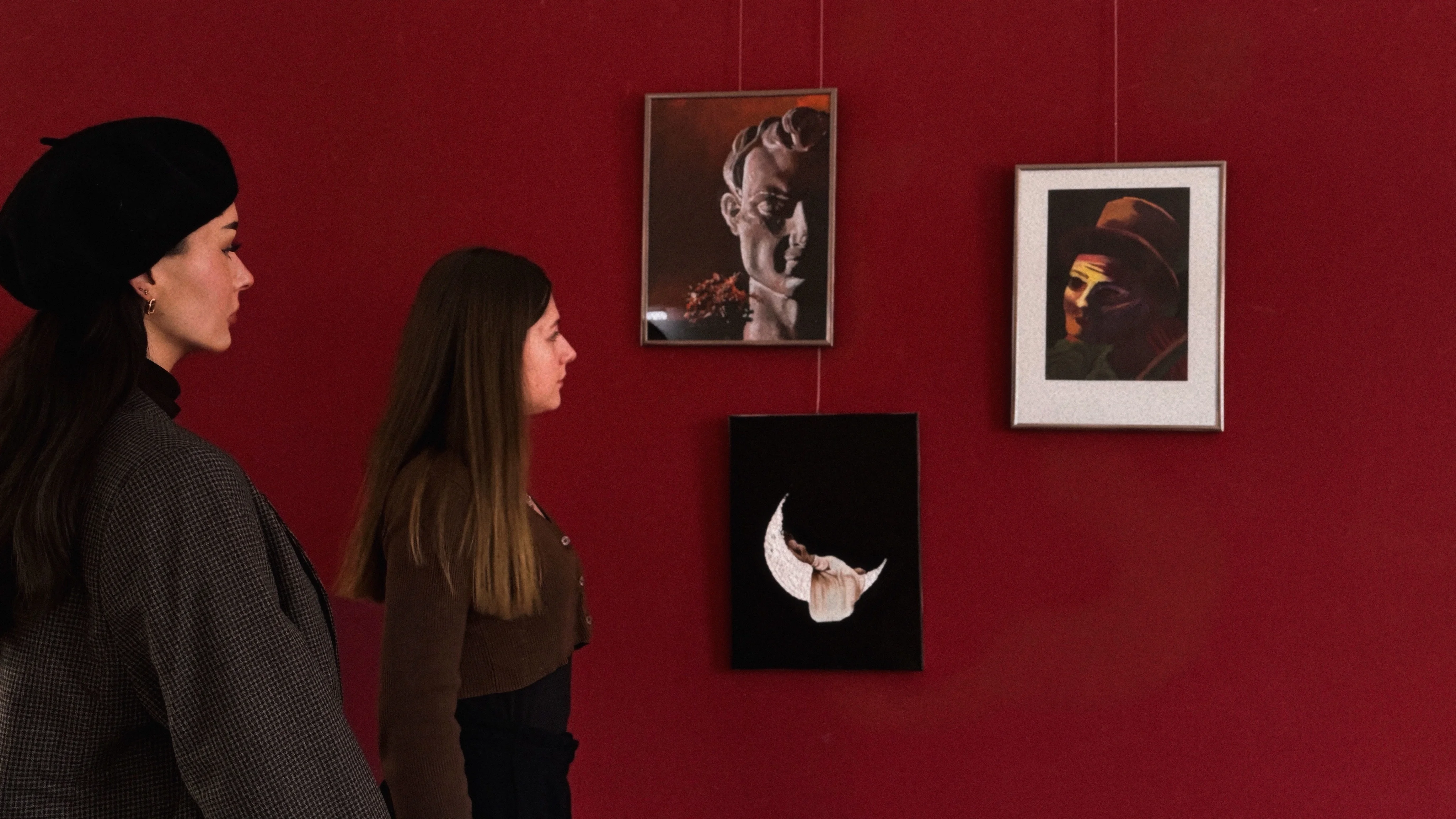Zwei Frauen betrachten drei an einer dunkelroten Wand hängende Kunstwerke in der Ausstellung "Young Visions" in Marienberg.