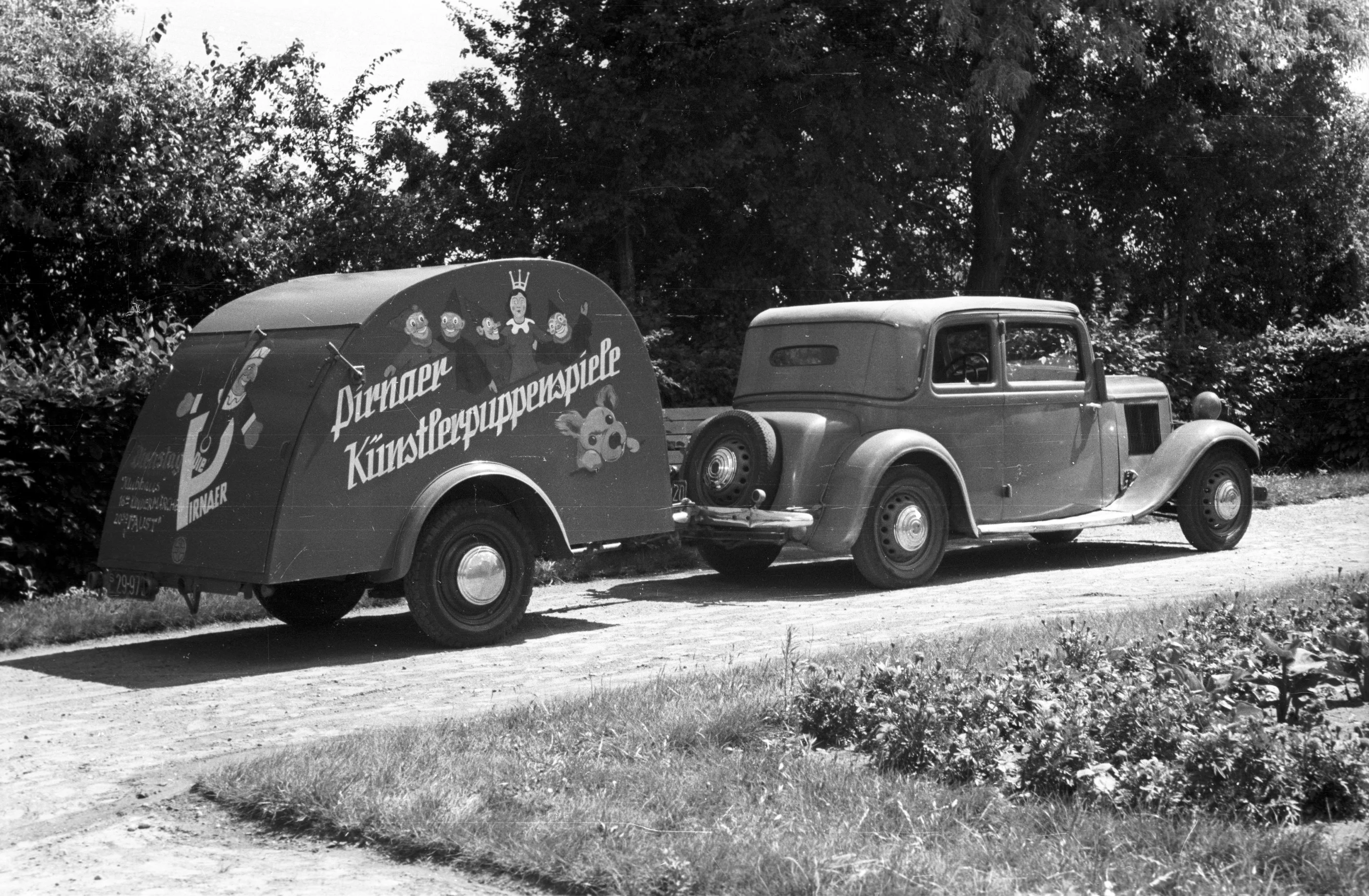 Historisches Schwarzweißfoto eines Oldtimer-Autos mit bemaltem Anhänger, auf dem "Pirnaer Künstlerpuppenspiele" steht