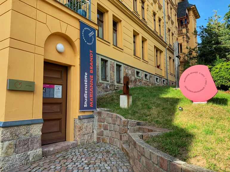 Detailansicht des Eingangsbereichs eines Gründerzeitgebäudes mit gelber Fassade, an der Tür sowie auf einer Grasfläche vor dem Haus Schilder mit Hinweisen zu den Marianne-Brandt-Studienräumen und zur Marianne-Brandt-Gesellschaft