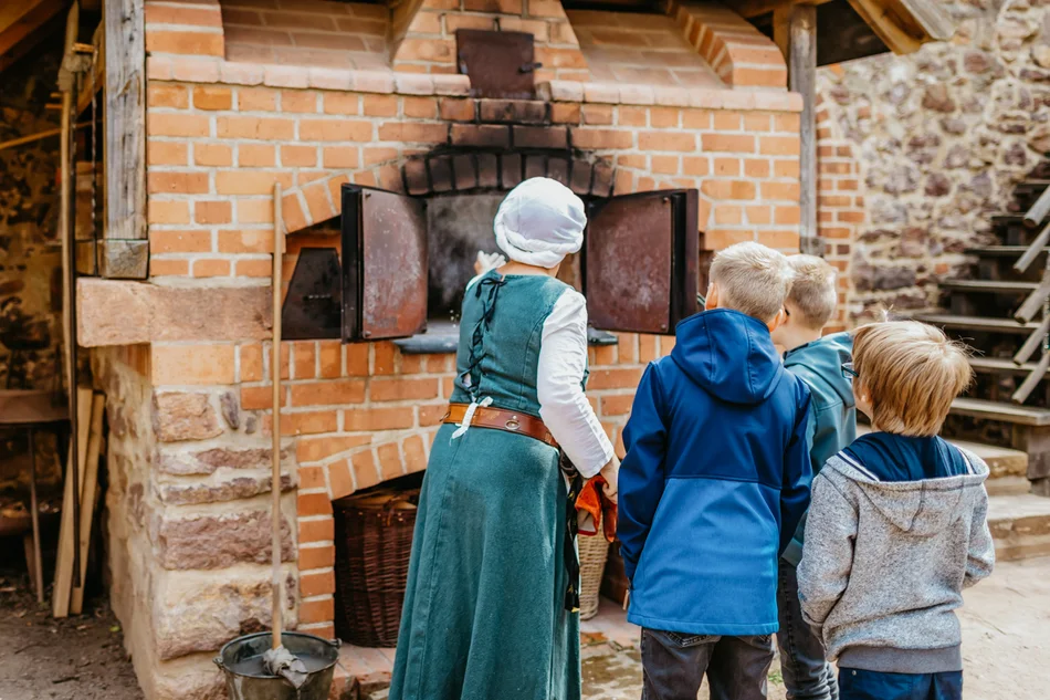 Eine Magd und einige Kinder schauen an der geöffneten Ofentür - ob die Temperatur schon reicht zum Backen?