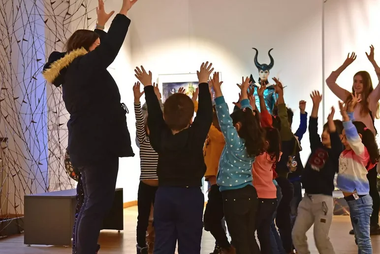 Angeleitet von zwei Frauen steht eine Kindergruppe in einem Ausstellungsraum; die Frauen und die Kinder strecken gemeinsam die Arme in die Hhe.