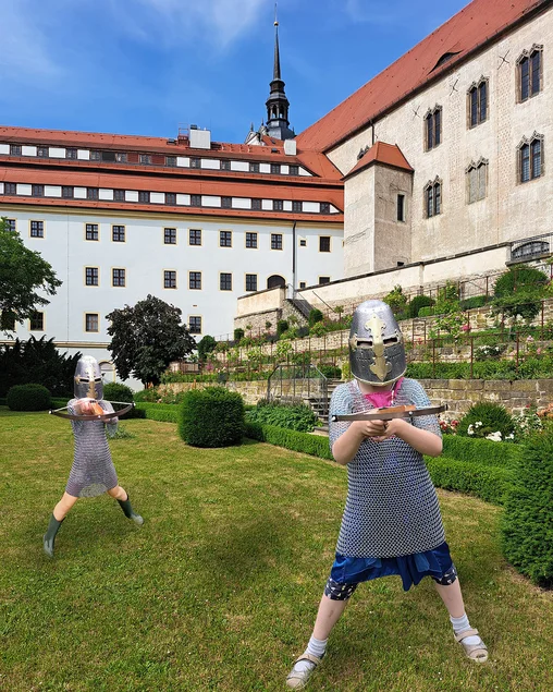 Zwei Mädchen mit Kettenhemden, Helmen und Armbrüsten stehen im Rosengarten von Schloss Hartenfels in Torgau.