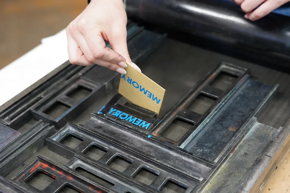Drucken an der Handpresse In einen Druckstock sind die das Wort MEMORY bildenden Blei-Lettern gespannt, diese sind mit blauer Farbe eingestrichen. Eine Hand löst davon ein kleines Papierkärtchen, auf dem das gedruckte Wort steht.
